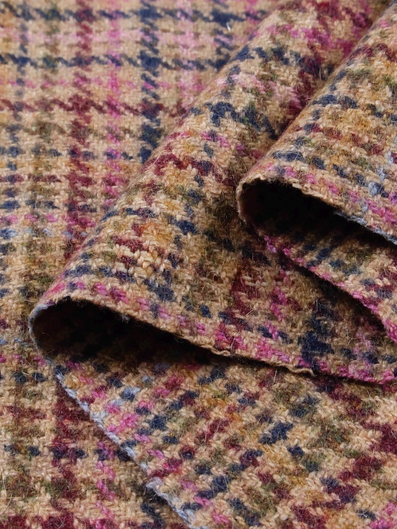 May include: Close-up of a folded fabric with a houndstooth pattern. The fabric features a blend of colors, including tan, pink, blue, and burgundy. The texture appears to be a woven tweed, suitable for apparel or home decor.
