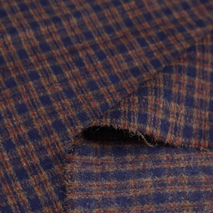 Op de afbeelding: Close-up van een blauw en bruin geruite stof. De stof heeft een getextureerd uiterlijk met een herhalend vierkant patroon. De kleuren zijn diepblauw en roodbruin, wat een klassieke, getextureerde uitstraling creëert. De stof is geschikt voor kleding of woondecoratie.