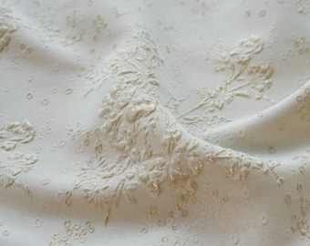 May include: Off-white fabric with a raised floral pattern and small, scattered dot details. The design features embossed flowers and foliage in a lighter shade, creating a textured, elegant look. The fabric is folded, showing the interplay of light and shadow.