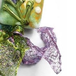 May include: A detailed view of a handmade artwork. It features a green and yellow glass component with white details, connected to a fine, web-like structure in purple and green hues, set against a white backdrop.