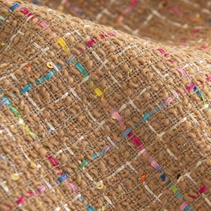 Op de afbeelding: Close-up van een textiel met een geweven patroon. De stof is voornamelijk bruin van kleur met witte en kleurrijke draden van roze, blauw, geel en groen die er doorheen zijn geweven. De stof heeft een licht ruwe, noppenachtige textuur.