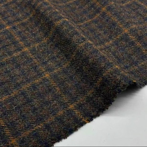 Op de afbeelding: Close-up van een textiel met een ruitpatroon. De stof is een mix van donkerbruine, marineblauwe en goudgele draden, wat een warme, rustieke uitstraling creëert. De randen van de stof zijn gerafeld.