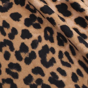 Puede incluir: Primer plano de una tela con estampado de leopardo. La tela tiene un fondo beige con manchas negras de varios tamaños y formas. La tela está doblada, mostrando la textura y el patrón.