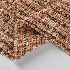 May include: A textured tweed fabric featuring a woven pattern of burnt orange, cream, and black threads. The material includes metallic gold fibres interspersed throughout the weave, creating a shimmering effect across the dense, multi-coloured textile.
