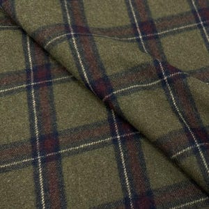 May include: Close-up of olive green plaid fabric with a traditional pattern. The fabric has a repeating grid of dark red, navy blue, and white lines. The material is folded, showing the texture and drape of the textile.