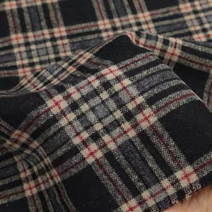 May include: Close-up of a folded fabric with a classic plaid pattern. The fabric features a dark navy blue base with intersecting lines in beige, grey, and thin red stripes. The material appears soft and textured, suitable for apparel or home decor.