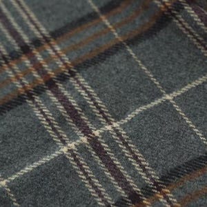 Op de afbeelding: Close-up van een grijze geruite stof met elkaar kruisende lijnen in wit, bruin en donkerpaars. De stof heeft een getextureerd uiterlijk, wat wijst op een geweven materiaal zoals wol of flanel. Het patroon is een klassiek tartan ontwerp.