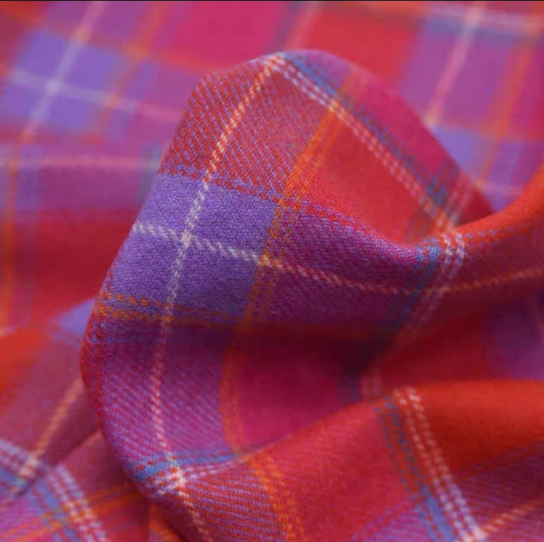 May include: A close-up shot of a colourful plaid fabric, predominantly red, pink, and purple. The tartan pattern is clearly visible, with intersecting lines creating a classic design. The fabric is folded, revealing its texture and depth.
