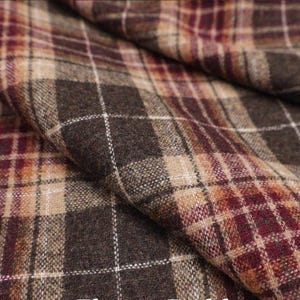 Può includere: Primo piano di un tessuto piegato con un motivo a quadri. Il tessuto presenta un mix di colori marrone, beige e bordeaux, con linee a griglia bianche. La trama appare morbida e tessuta, suggerendo un potenziale per abbigliamento o arredamento.