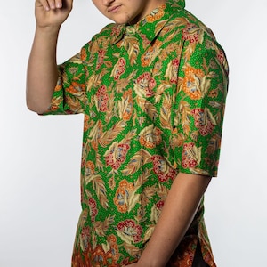 Op de afbeelding: Groen overhemd met een bloemenpatroon in rood, oranje en geel. Het overhemd heeft korte mouwen en een kraag. Een zwarte vissershoed met witte tekst wordt gedragen. Het overhemd wordt gecombineerd met een grijze broek.