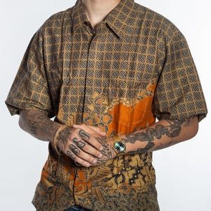 Op de afbeelding: Korte mouwen overhemd met een geometrisch patroon in bruin en grijs. Het overhemd heeft een oranje paneel met een bloemenmotief. Het overhemd is gemaakt van een lichte stof.