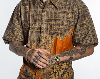 May include: Short-sleeved shirt with a geometric pattern in brown and grey. The shirt has an orange panel with a floral design. The shirt is made of a lightweight fabric.