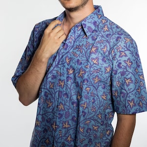 Puede incluir: Camisa de manga corta azul claro, adornada con un estampado floral repetido en tonos morados y beige. La camisa presenta un cuello clásico y un corte relajado, adecuada para el uso diario.