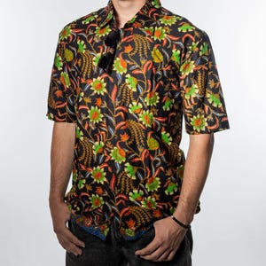 Puede incluir: Camisa negra de manga corta con un vibrante estampado floral en verde, naranja y amarillo. La camisa tiene un cuello clásico y se combina con vaqueros oscuros. Gafas de sol en el cuello.