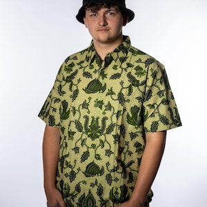 Op de afbeelding: Een persoon draagt een zwarte vissershoed en een overhemd met korte mouwen en knopen. Het shirt heeft een groen en geel bloemenpatroon. De persoon draagt ook een grijze broek.