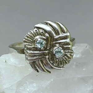 May include: A silver ring with a textured, abstract design featuring two clear gemstones. The ring is set on a white background.