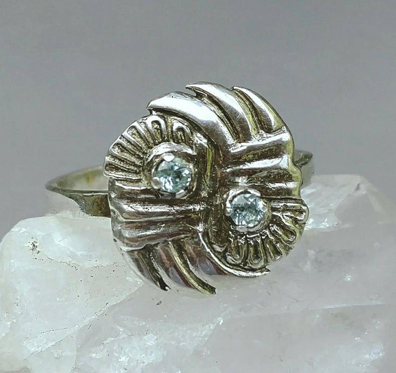 May include: A silver ring with a textured, abstract design featuring two clear gemstones. The ring is set on a white background.