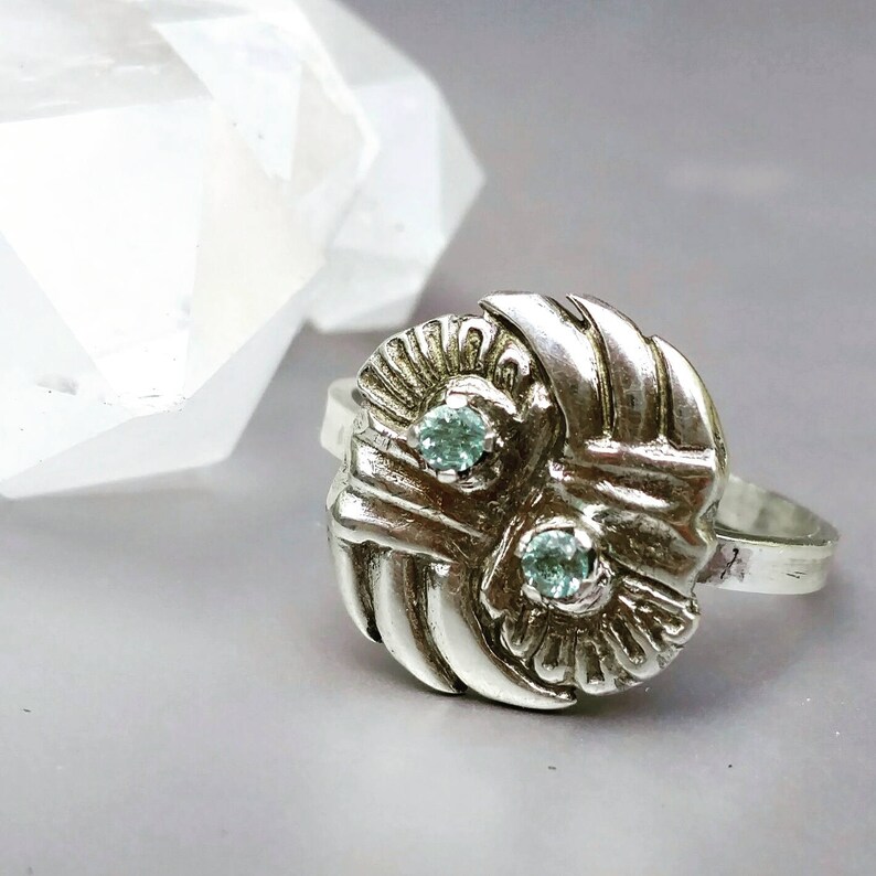 May include: A silver ring with a textured, circular design featuring two light blue gemstones. The ring is set on a white background.