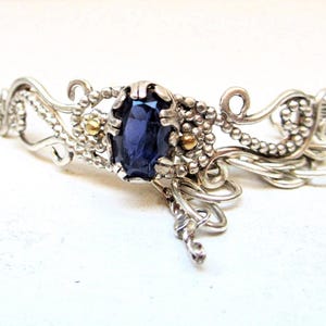 Può includere: Un bracciale argentato con una gemma ovale blu scuro al centro. Il bracciale presenta un'intricata lavorazione del metallo a spirale con piccoli accenti dorati e delicati dettagli floreali, creando un design elegante e ornato.