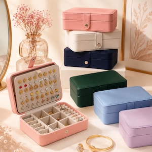 May include: A collection of rectangular jewelry boxes in pink, white, navy blue, dark green, light blue, and lavender. One open pink box displays a beige interior with earring holes and divided compartments. Gold rings and a bracelet are placed nearby.
