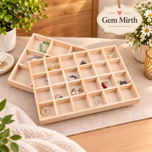 May include: A light-colored wooden jewelry organizer tray featuring a grid of square compartments. The tray contains various small earrings, rings, and a beaded bracelet. A hanging sign above displays the text Gem Mirth with a small heart icon.