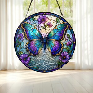Op de afbeelding: Een ronde glas-in-lood zonnevanger met een grote blauwe en paarse vlinder in het midden, omringd door paarse bloemen en groene ranken. De tekst A Beautiful New Chapter staat in witte sierletters onderaan het glazen paneel.