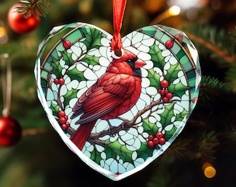May include: Heart-shaped Christmas ornament with a stained-glass effect, depicting a red cardinal bird on a branch with holly and berries. The ornament hangs from a red ribbon. The background is a blurred Christmas tree.