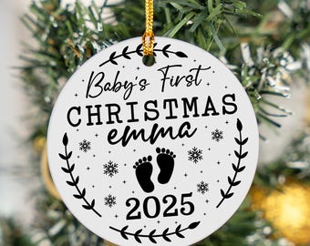 Puede incluir: Adorno navideño de cerámica blanca con cordón dorado para colgar. El adorno presenta el texto "Baby's First Christmas Emma 2025" con huellas de pies negras y diseños de copos de nieve. Un borde decorativo de hojas rodea el texto.