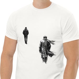 Peut inclure: T-shirt blanc avec une illustration en noir et blanc de deux personnages s'éloignant. L'un porte un manteau et une écharpe, l'autre une veste. Le t-shirt est fait d'une matière douce et confortable.