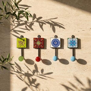 Può includere: Quattro ganci da parete con piastrelle decorative quadrate dai motivi distinti. Il primo presenta un cammello verde, il secondo un motivo geometrico rosso, il terzo un mandala blu e il quarto un motivo floreale blu, ciascuno con una sfera colorata.