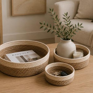 May include: Three woven rope baskets of varying sizes on a wooden table. The largest basket holds a book titled "KINFOLK." The middle basket contains a vase with greenery and a candle. The smallest basket holds keys.