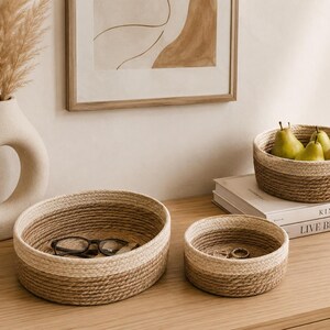 May include: A neutral-toned interior decor arrangement featuring woven rope baskets, a decorative vase, and a framed artwork. The baskets are in varying sizes, with one holding eyeglasses and another holding jewelry. Pears are in a basket on top of books.