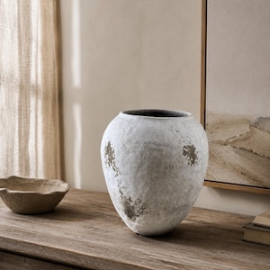 May include: A large, rounded ceramic vase with a textured, weathered white finish featuring patches of dark grey and brown distressing. The vessel has a wide, open mouth and a tapered base, showcasing a rough, uneven surface throughout its exterior.
