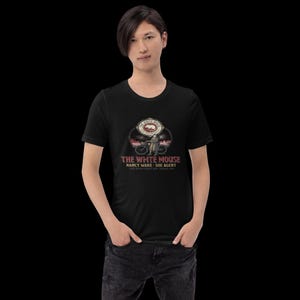 Pode incluir: Uma t-shirt preta de manga curta com uma impressão gráfica de uma pessoa com uma bicicleta. O texto diz THE WHITE MOUSE, NANCY WAKE - SOE AGENT, THEY NEVER CAUGHT HER - FRANCE 1944. O design inclui um emblema circular com uma ilustração de um rato.