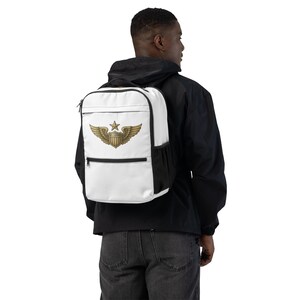 Pode incluir: Mochila branca com acabamento preto, com um emblema dourado com uma estrela, asas e um escudo. A mochila tem um bolso frontal com zíper e um bolso lateral. A mochila está sendo usada por uma pessoa.