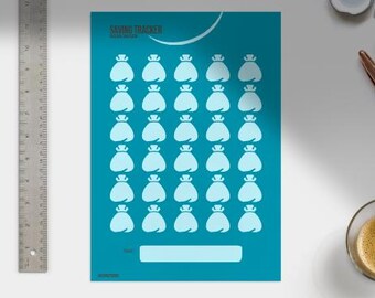 May include: Teal-colored savings tracker with white illustrations of money bags. The text "SAVING TRACKER" is at the top. A metal ruler is on the left. A pen and a cup of coffee are on the right.