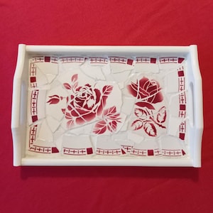 Può includere: Vassoio rettangolare bianco con un design a mosaico. Il vassoio ha una cornice e manici bianchi. Il mosaico è composto da piastrelle bianche rotte con disegni e accenti di rose rosse. Il vassoio è su sfondo rosso.