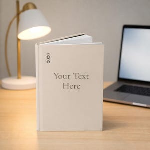 Könnte beinhalten: Ein beigefarbenes Hardcover-Buch mit dem Text "Your Text Here" und dem Jahr "2026" auf dem Einband. Das Buch ist geöffnet, mit weißen Seiten sichtbar. Eine Schreibtischlampe und ein Laptop sind im Hintergrund.