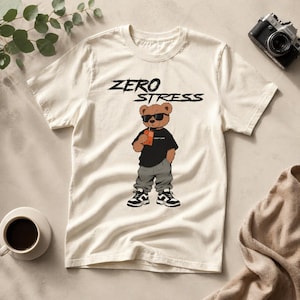 Könnte beinhalten: Cremefarbenes T-Shirt mit dem Text "ZERO STRESS" über einem Cartoon-Bären mit Sonnenbrille, schwarzem Shirt, grauer Hose und Turnschuhen. Der Bär hält ein Getränk. Eine Kamera und eine Kaffeetasse befinden sich im Hintergrund.