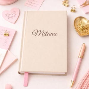 Könnte beinhalten: Ein beiges Notizbuch mit dem Namen "Milana" in Schreibschrift, ein rosa Stift und ein rosa Lesezeichen. Das Bild enthält auch eine rosa Kaffeetasse, eine herzförmige Schale mit goldenen Büroklammern und andere rosa Schreibwaren.