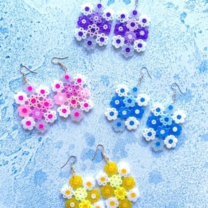 May include: A selection of handmade earrings in various colours. The earrings are made of small, colourful beads arranged in a floral pattern. Colours include purple, pink, blue, and yellow. Each pair has a gold-coloured hook.