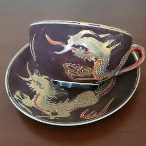 Op de afbeelding: Een donkerbruine theekop en schotel met een gouden en rood drakenontwerp. De kop en schotel zijn versierd met een gouden rand.