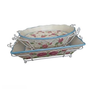 May include: Two ceramic baking dishes with a cream-colored base, featuring a pink rose floral pattern and a light blue rim. Each dish is set within a white metal wire rack with decorative scrollwork handles and small ball-shaped feet.