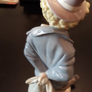 May include: A porcelain figurine of a clown with blonde hair wearing a blue jacket and a hat with a white band. The clown is holding a white bag in their right hand.