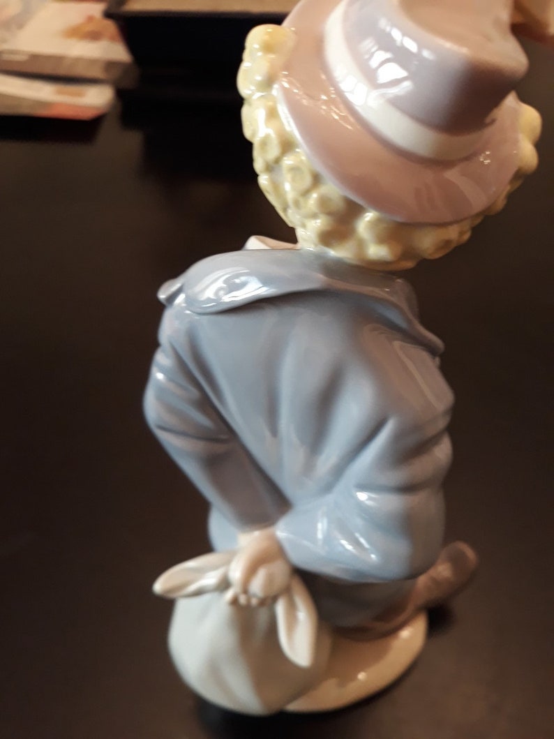 May include: A porcelain figurine of a clown with blonde hair wearing a blue jacket and a hat with a white band. The clown is holding a white bag in their right hand.