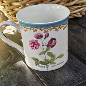 Op de afbeelding: Een witte keramische mok met een blauw en gouden bloemenontwerp. De mok heeft een roze roos met groene bladeren en de tekst "Rosa Indica dichotoma".