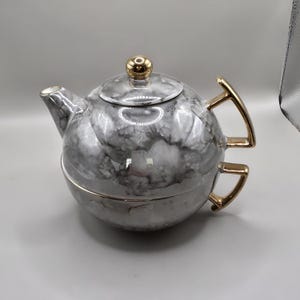 May include: A round ceramic teapot and teacup set featuring a gray and white marbled pattern. The teapot has a gold-colored lid knob and a gold-colored handle. The teacup, which nests beneath the teapot, also features a gold-colored handle.