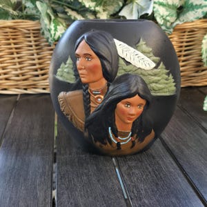 May include: A black ceramic vase with a relief design featuring two Native American women. The women have long black hair and wear necklaces. A white feather and green trees are also depicted. The vase is on a dark wooden surface.