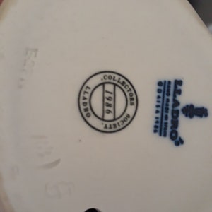 May include: White porcelain figurine with a blue and white Lladro logo and a black circle with the text "Collectors Society 1986" on the bottom.