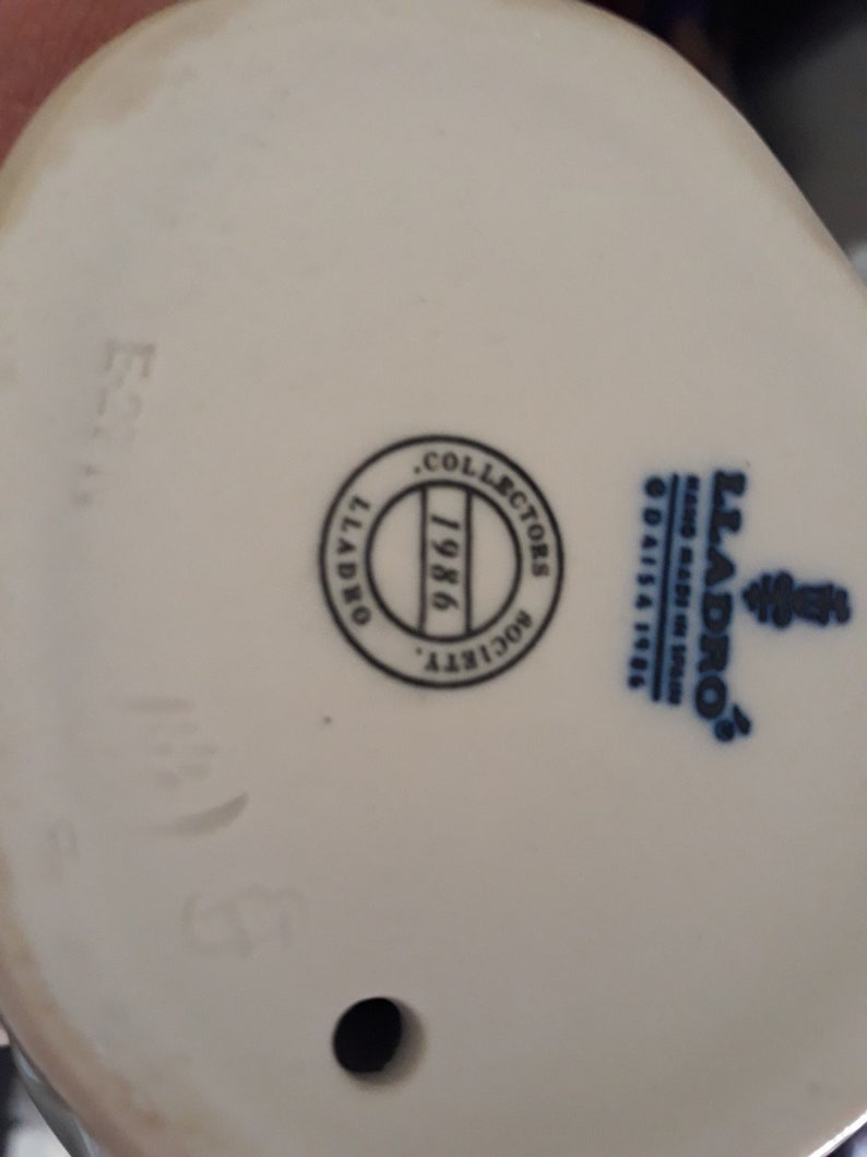 May include: White porcelain figurine with a blue and white Lladro logo and a black circle with the text "Collectors Society 1986" on the bottom.